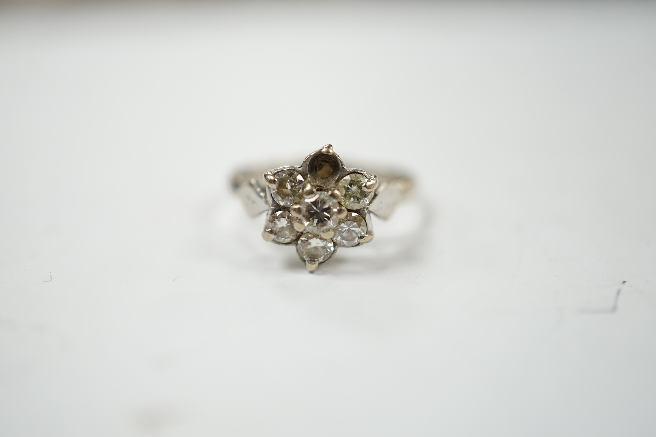 An 18ct white metal and seven stone diamond cluster set flower head ring (one stone missing), size K/L, gross weight 3.4 grams.
